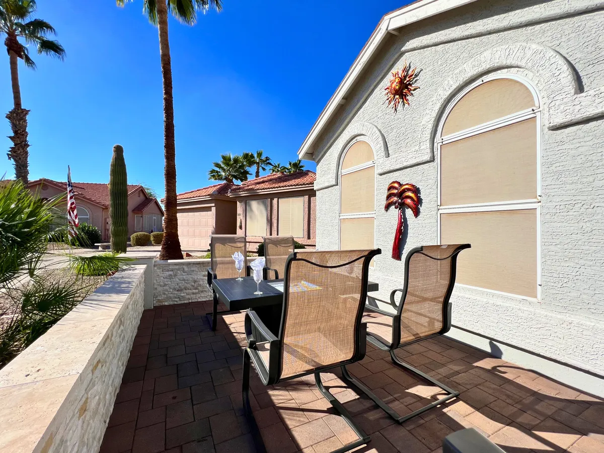 A stylish patio seating area is surrounded by decorative wall art and framed by tall palm trees, creating a welcoming outdoor living space. The setup includes comfortable chairs and a table, inviting visitors to dine or relax in the sun. The overall atmosphere feels relaxed and perfect for hosting gatherings.