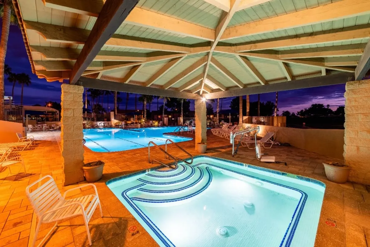 This image features a vibrant pool area with a jacuzzi, ample outdoor seating, and soft landscape lighting. The scene is set for socializing and relaxation, with several guests potentially gathering by the water and enjoying the tranquil evening ambiance.