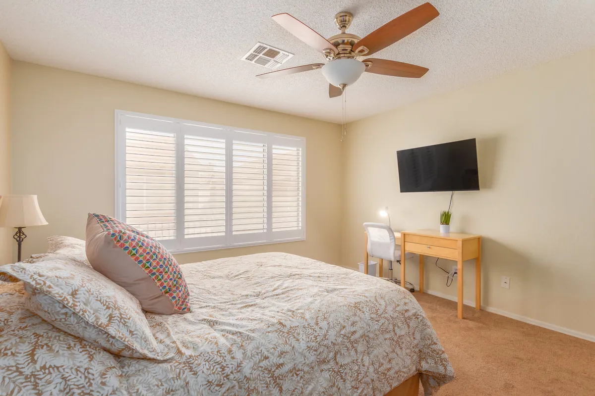 A serene bedroom setting with a bed, window shutters letting in gentle natural light, a TV for relaxing evenings, and a desk with a chair for correspondence or remote work. The room feels cozy and private, ideal for a restful retreat.