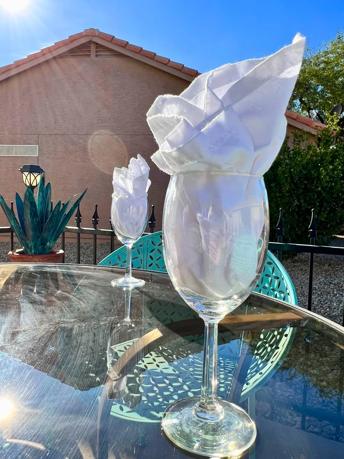 A beautifully set outdoor dining table with elegantly folded napkins in wine glasses, teal chairs, and a reflective glass tabletop. The table is framed by greenery and a bright, cheerful atmosphere, ideal for alfresco meals and gatherings.