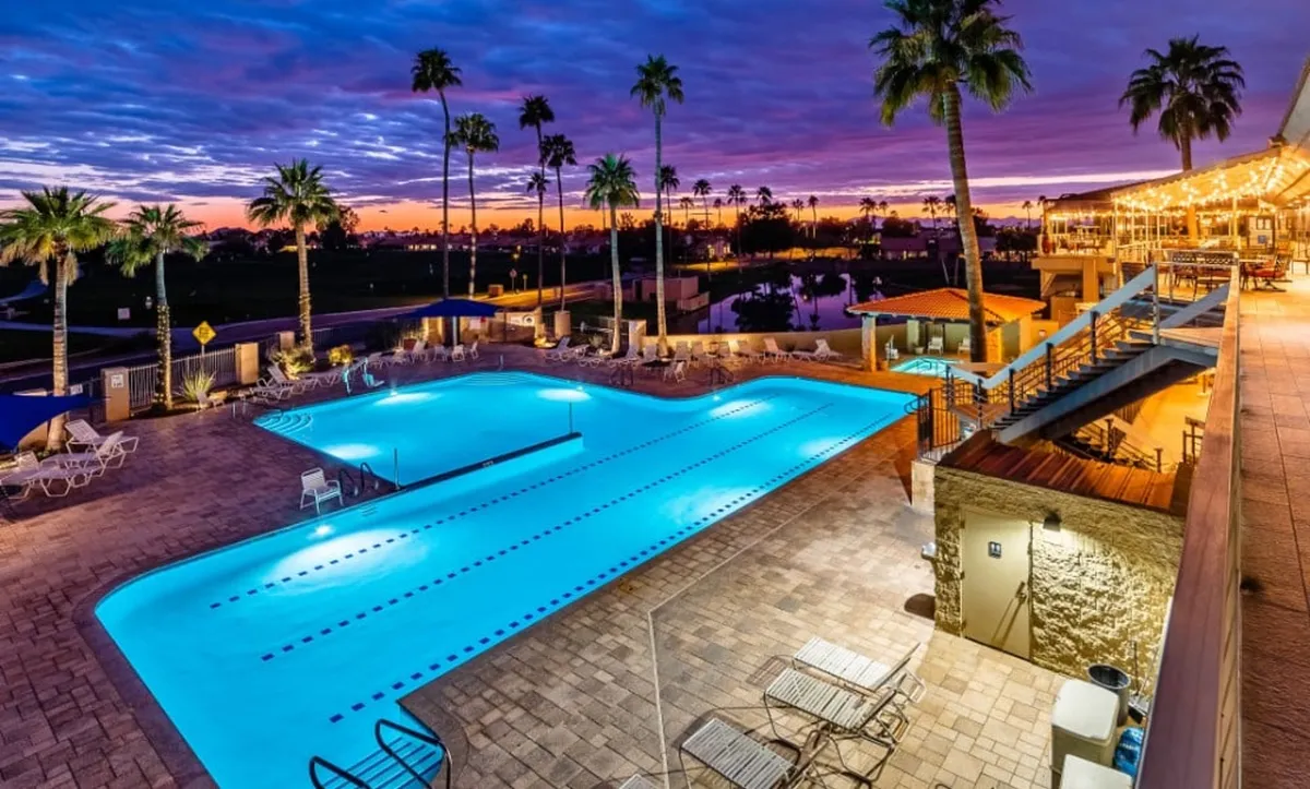 An expansive evening view of the resort's swimming pool, flanked by tall palm trees and vibrant sunset skies. The deck area offers spaces for sunbathing, social gatherings, and relaxation, all underscored by a lively yet serene ambiance.