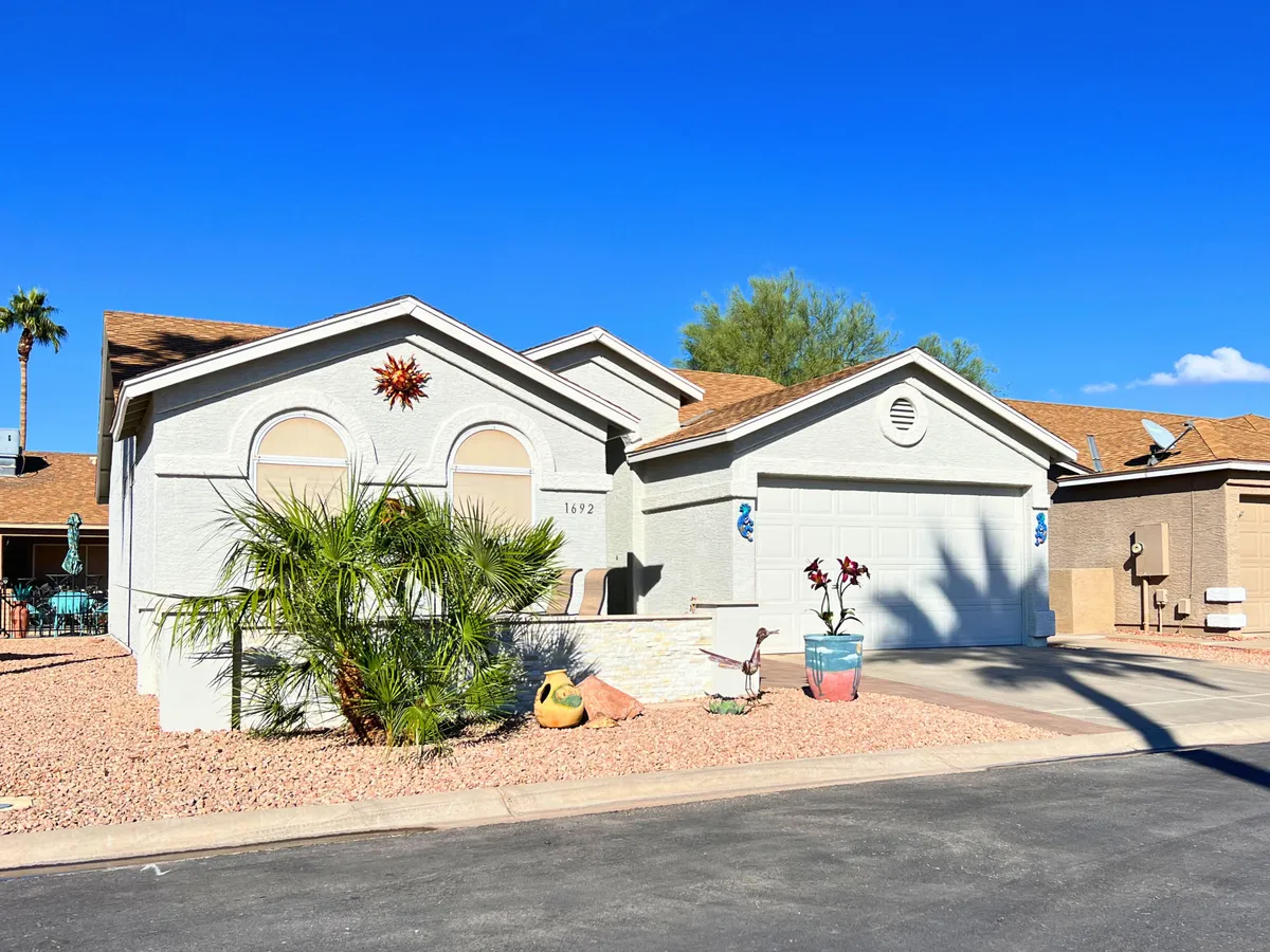 A charming house facade framed by a tall palm tree, colorful potted plants, and a decorative sun emblem. The porch area is welcoming and suggests a relaxed, sunny lifestyle, indicative of the Chandler and East Valley region.