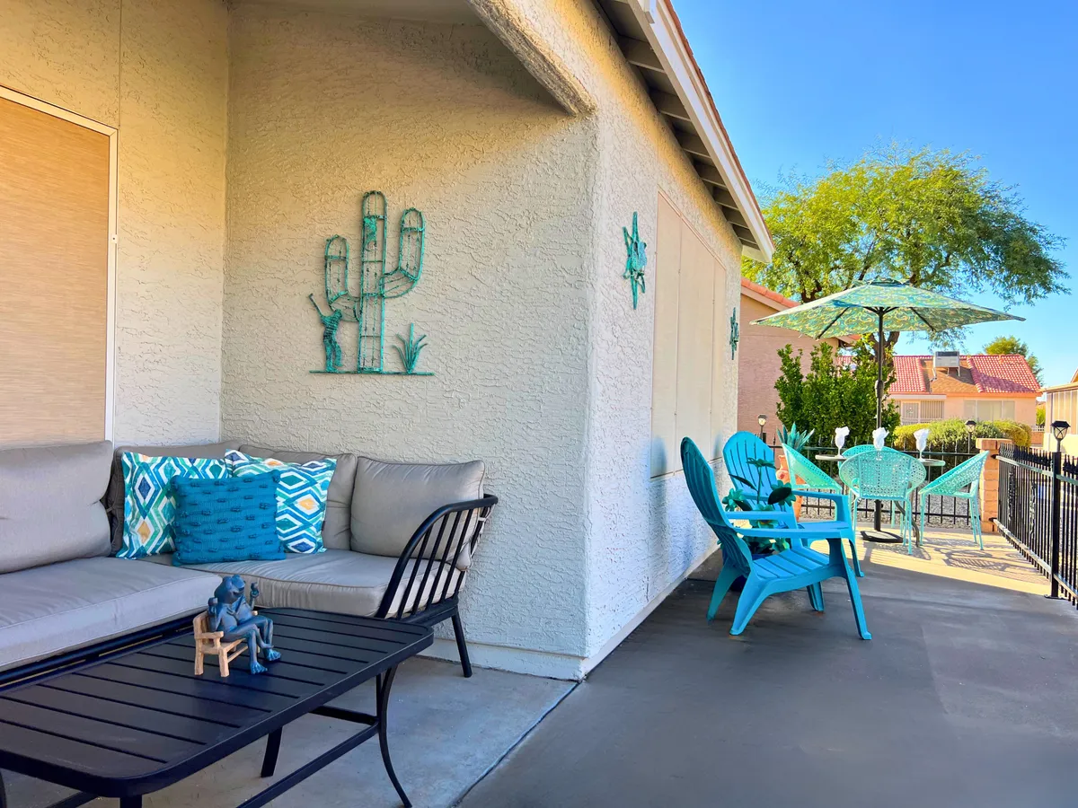 This image showcases a cozy outdoor seating area with blue cushioned chairs, decorative cactus wall art, and an umbrella providing shade. Lush greenery surrounds the space, creating an inviting atmosphere for relaxation or social gatherings at sunset.