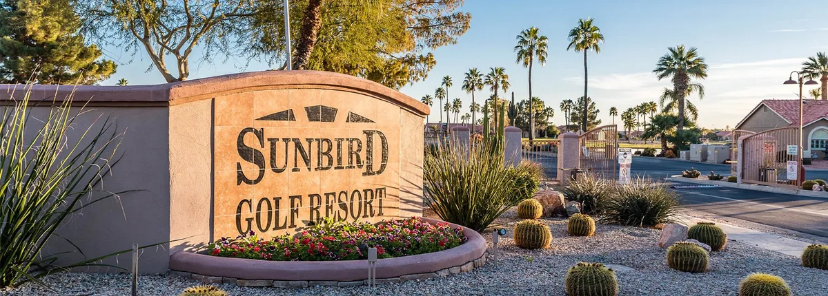 This exterior image highlights the welcoming entrance signage for the 'Sunbird Golf Resort,' surrounded by well-maintained landscaped flower beds, palm trees, and cactus plants. The setting conveys the inviting atmosphere and pride in the resort community.