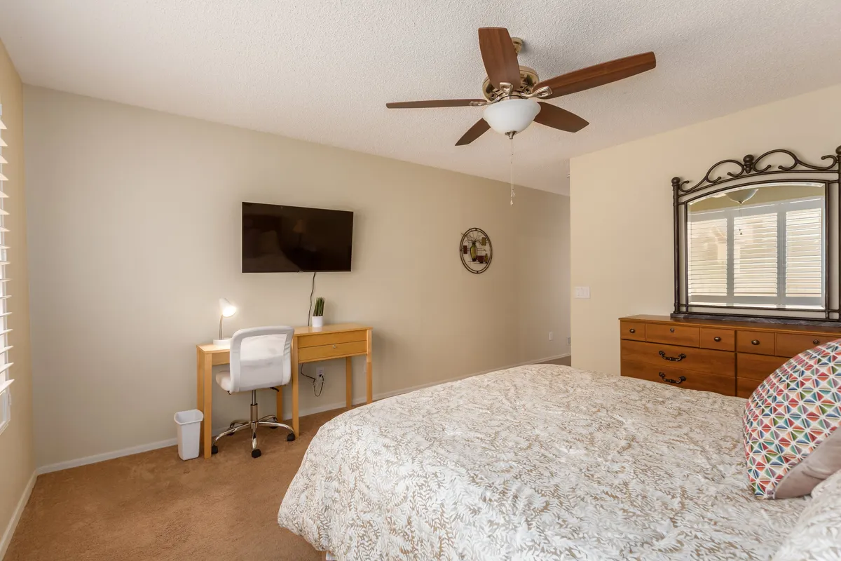 This image features a cozy bedroom with a queen bed, a desk positioned for productive work sessions, a television for personal entertainment, and a mirror. The room appears calm and comfortable, with plenty of natural light filtering in, making it ideal for unwinding after a busy day.